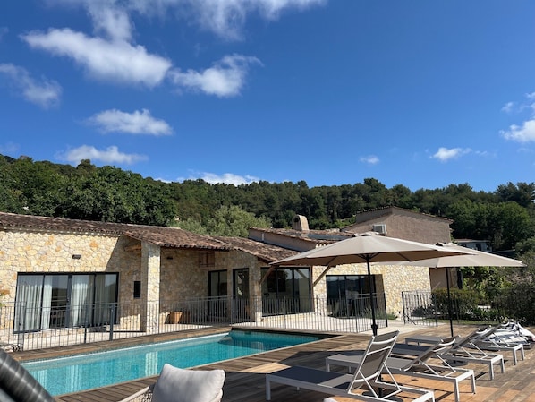 A poolside area with lounge chairs and umbrellas, a stone building with a tiled roof, and a forested hillside in the background.