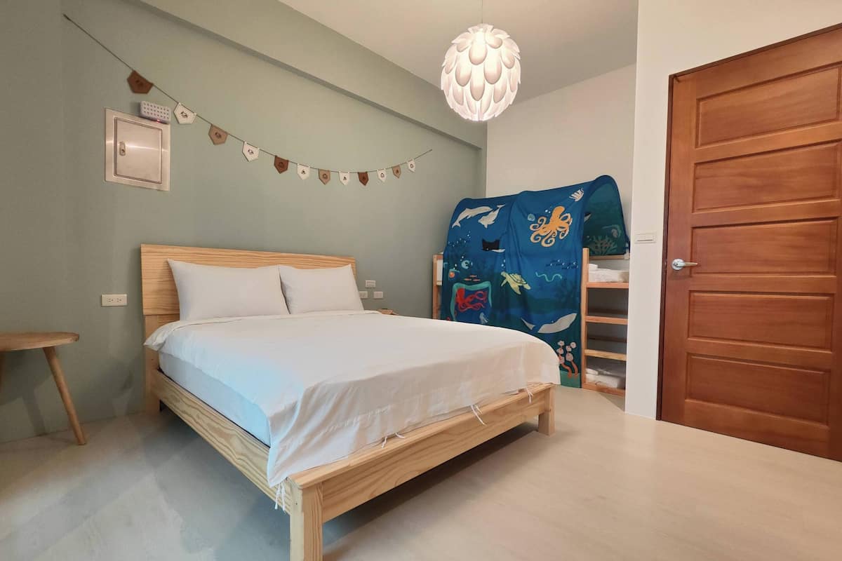 Quarto com cama de madeira, luminária pendente, tomada na parede e uma porta.