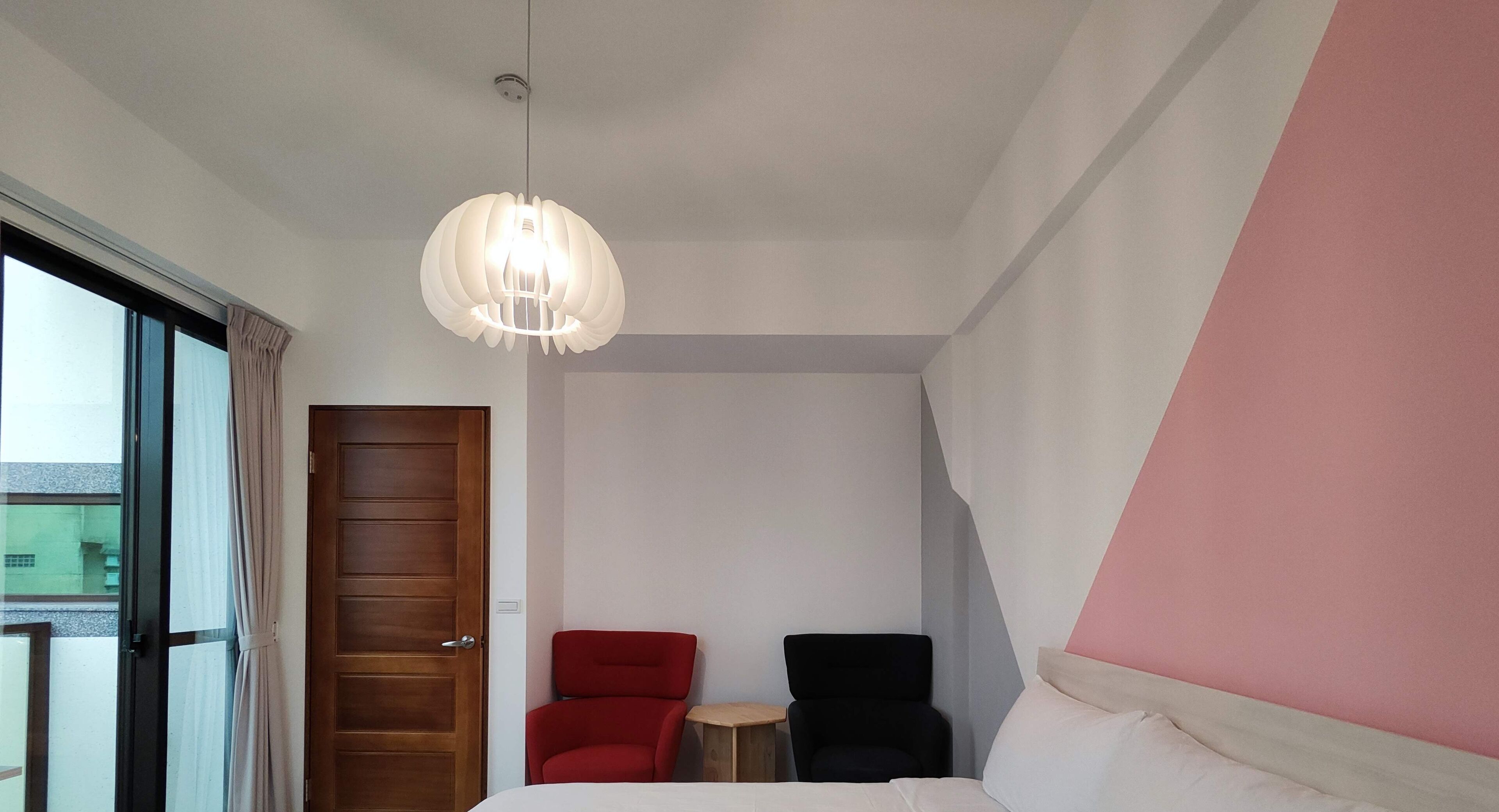 A hotel room with a bed, a red armchair, a black chair, a small table, and a large window.