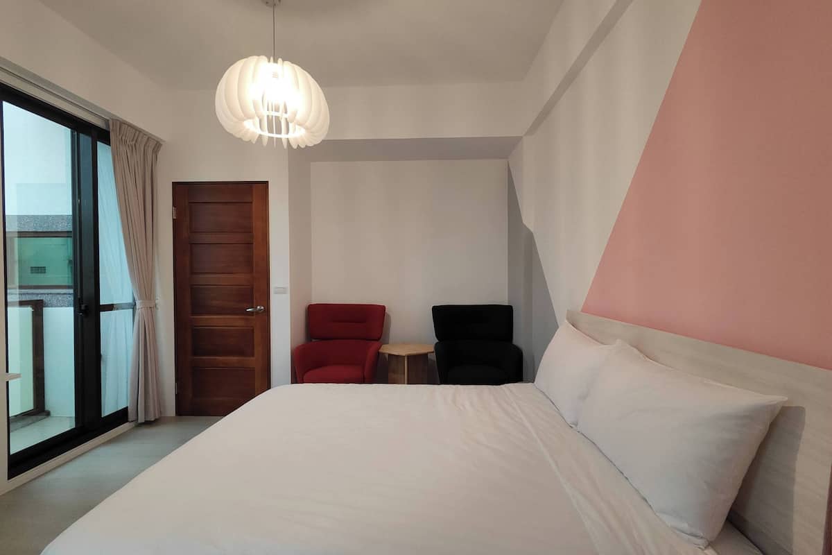 Quarto de hotel com uma cama grande, uma cadeira vermelha, uma cadeira preta, uma mesa pequena e uma janela ampla com cortinas.