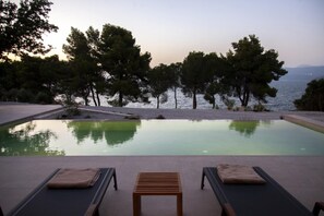 Una piscina con due lettini e un tavolo di legno, circondata da alberi e vista sul mare.
