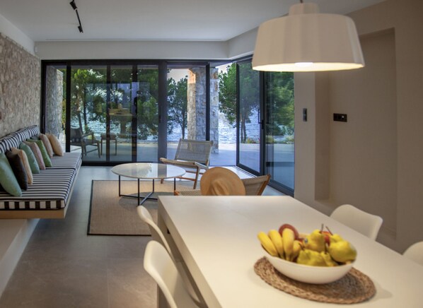 A modern living room with a sofa, dining table, and chairs. There is a bowl of fruit on the table. - Theros Estate 3- 3 BED (Amfilochia)