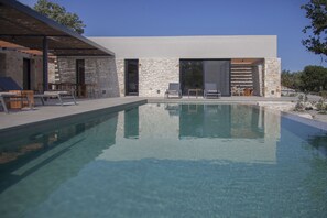 A modern pool area with clear water, stone walls, and outdoor seating. - Theros Estate 3- 3 BED (Amfilochia)