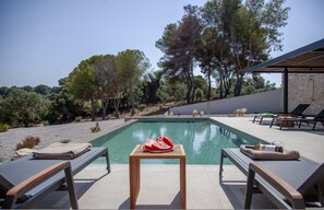 A pool area with lounge chairs, a table with a watermelon, and a stone wall. - Theros Estate 3- 3 BED (Amfilochia)