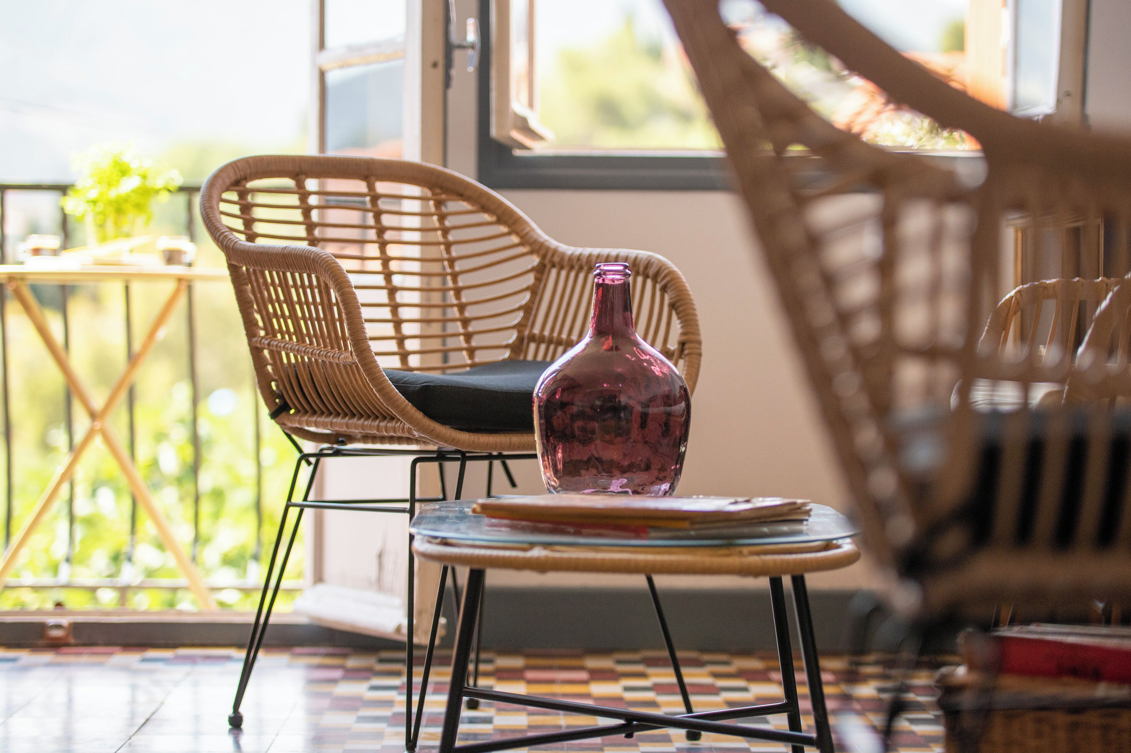 A wicker chair with a cushion, a small round table with a bottle, and a patterned floor.