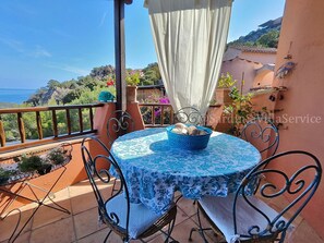 Ein runder Tisch mit blumigem, blauem Tischläufer, umgeben von vier Metallstühlen, auf einem Balkon mit Blick aufs Meer.