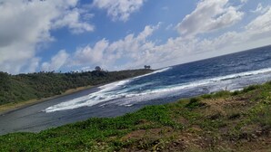 Eine Küstenlandschaft mit blauem Himmel, Wellen, die auf einen Sandstrand treffen, und hügeligem Gelände mit Vegetation.