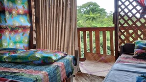 Ein Zimmer mit zwei Betten, jeweils mit bunt gemusterten Bettdecken. Ein Holzziergitter trennt den Raum von einem üppigen grünen Garten ab.