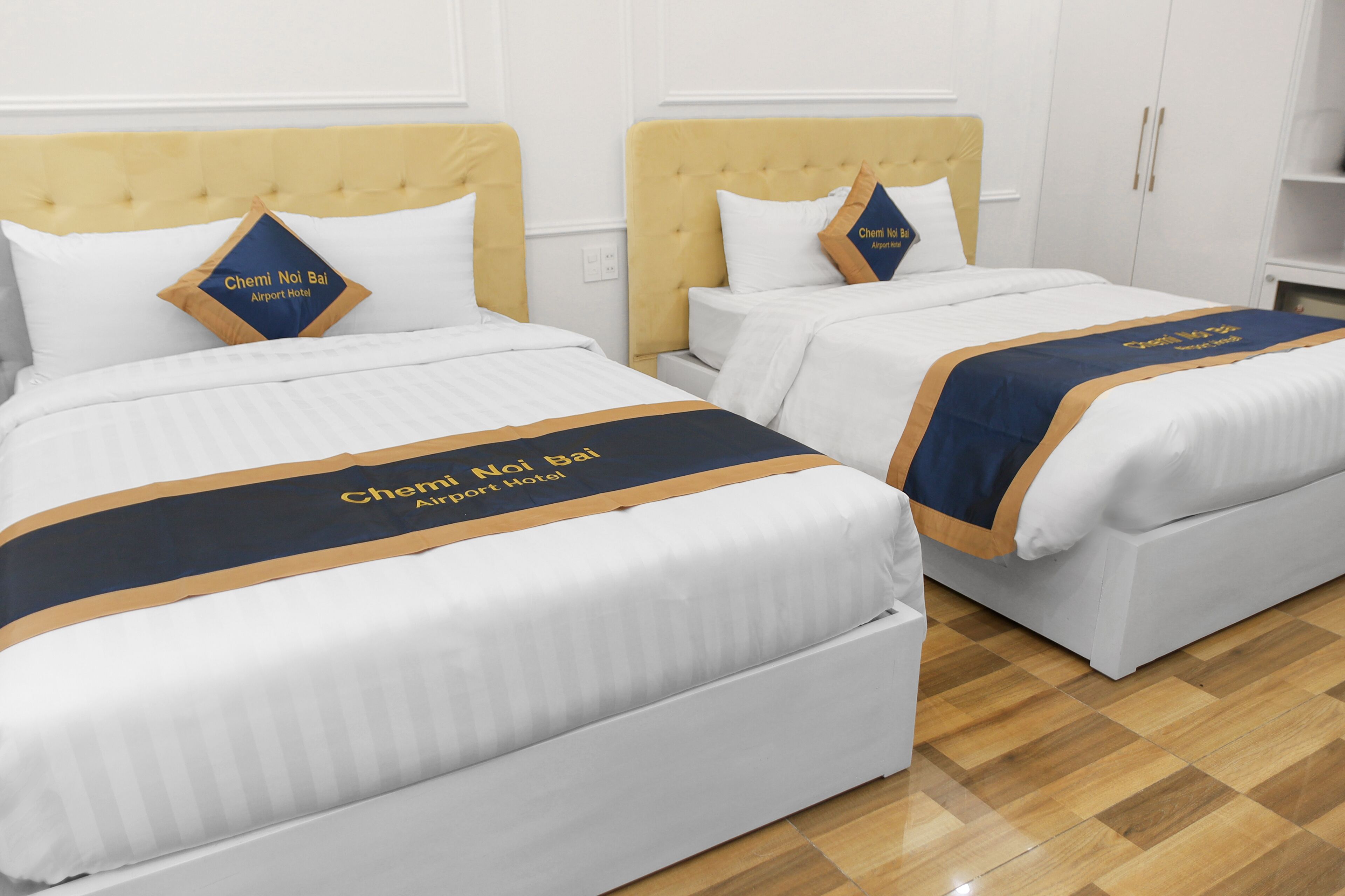 Zwei Betten mit weißer Bettwäsche und einem blau-goldenen Bettüberwurf. Jedes Bett hat ein Kopfkissen mit dem Text "Chemi Noi Bal Airport Hotel".