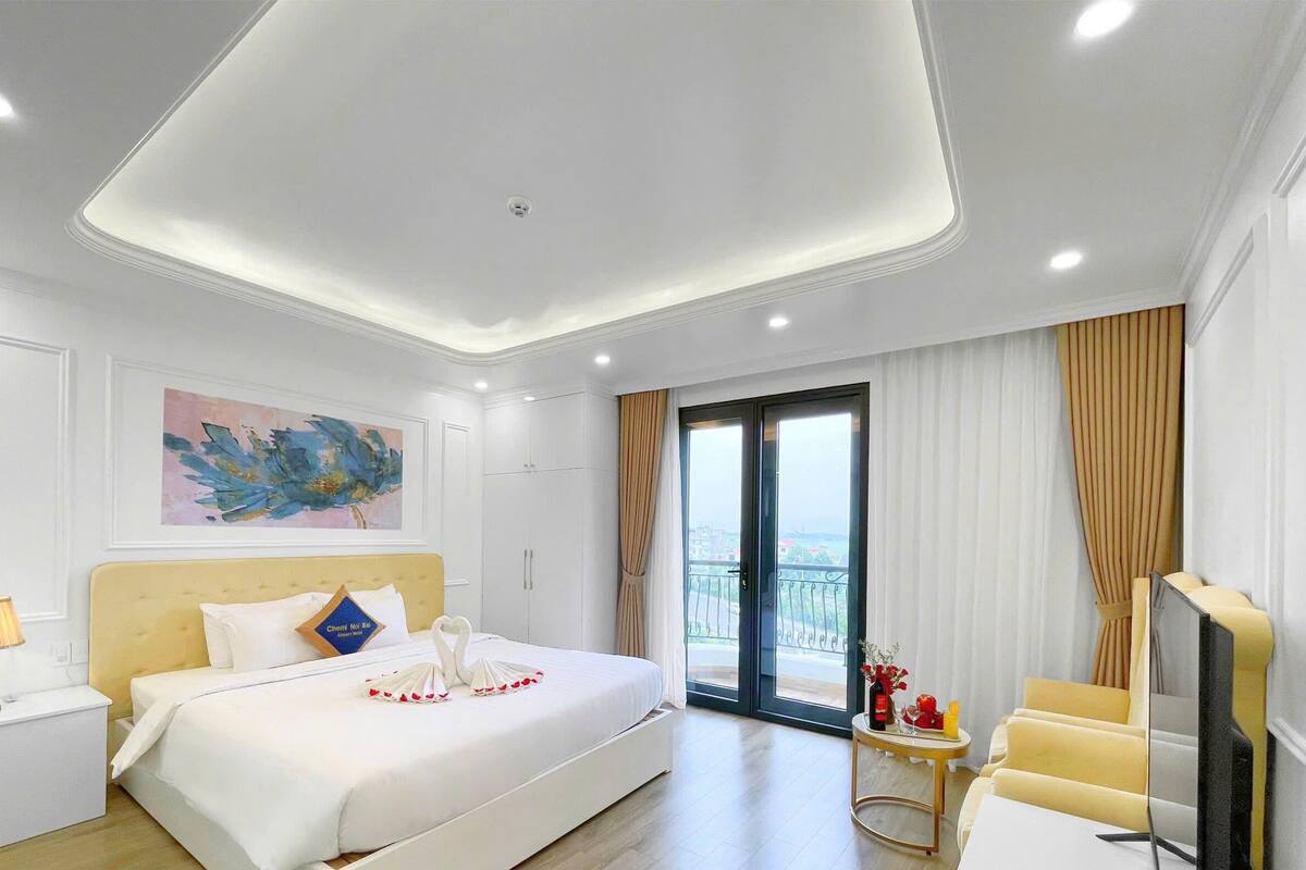 Habitación de hotel con cama, escritorio, silla, televisor y balcón con cortinas.