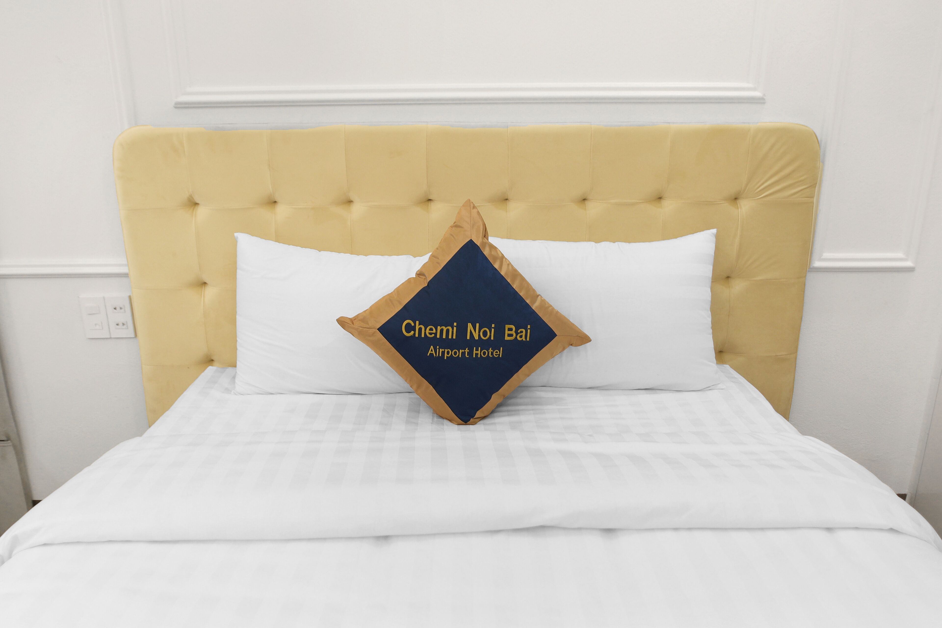 A bed with a tufted headboard, a white bedspread, and a pillow with "Chemi Noi Bai Airport Hotel" text.