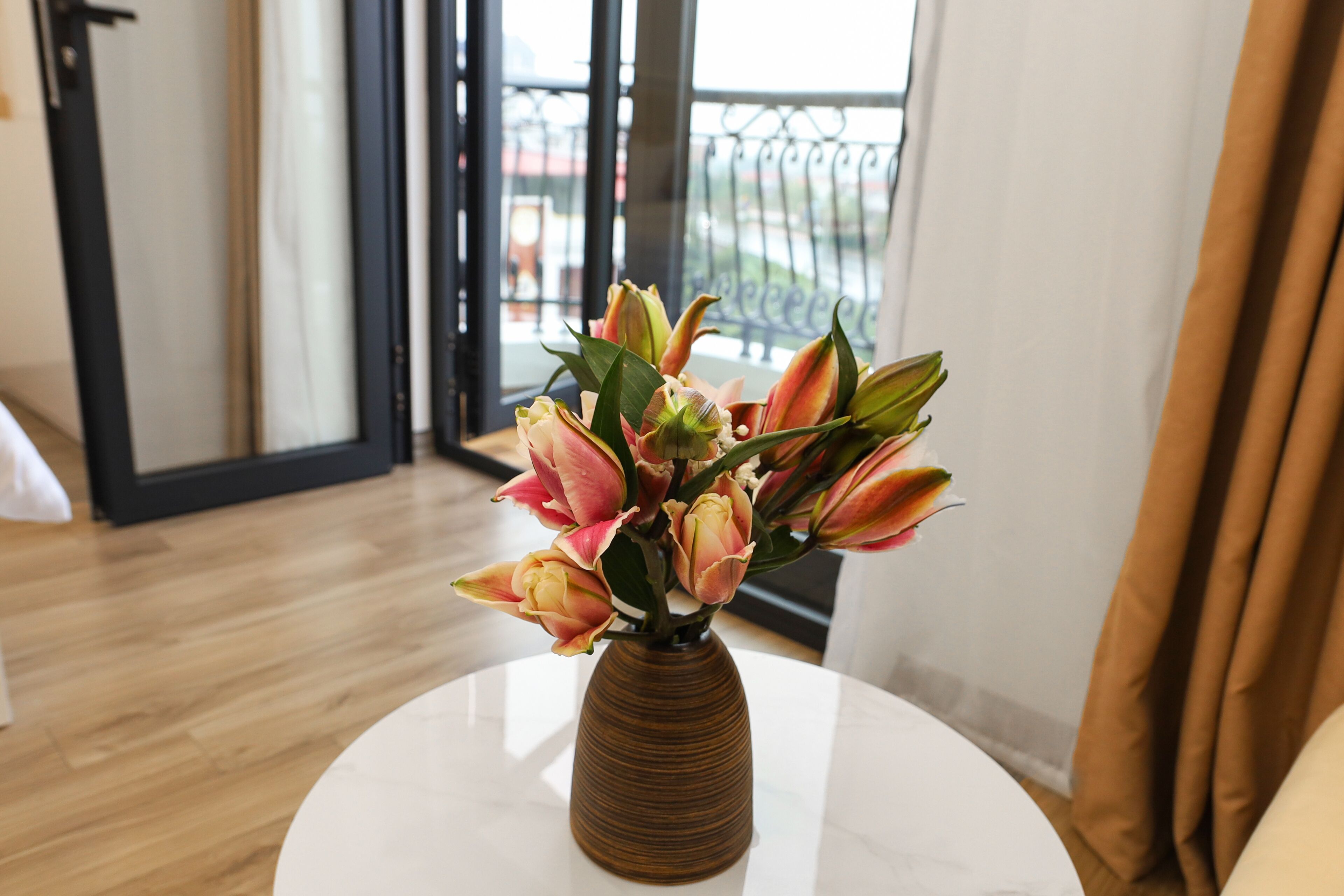 Eine Vase mit Blumen auf einem weißen Tisch in einem Raum mit Holzboden und Blick auf einen Balkon durch eine Glastür.