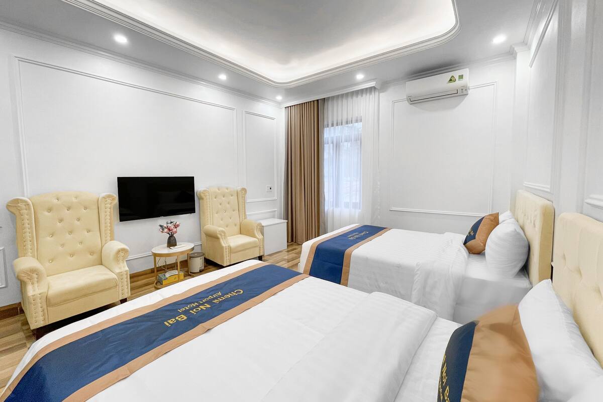 Habitación de hotel con dos camas, televisión, una mesita con un florero y ventana con cortinas.