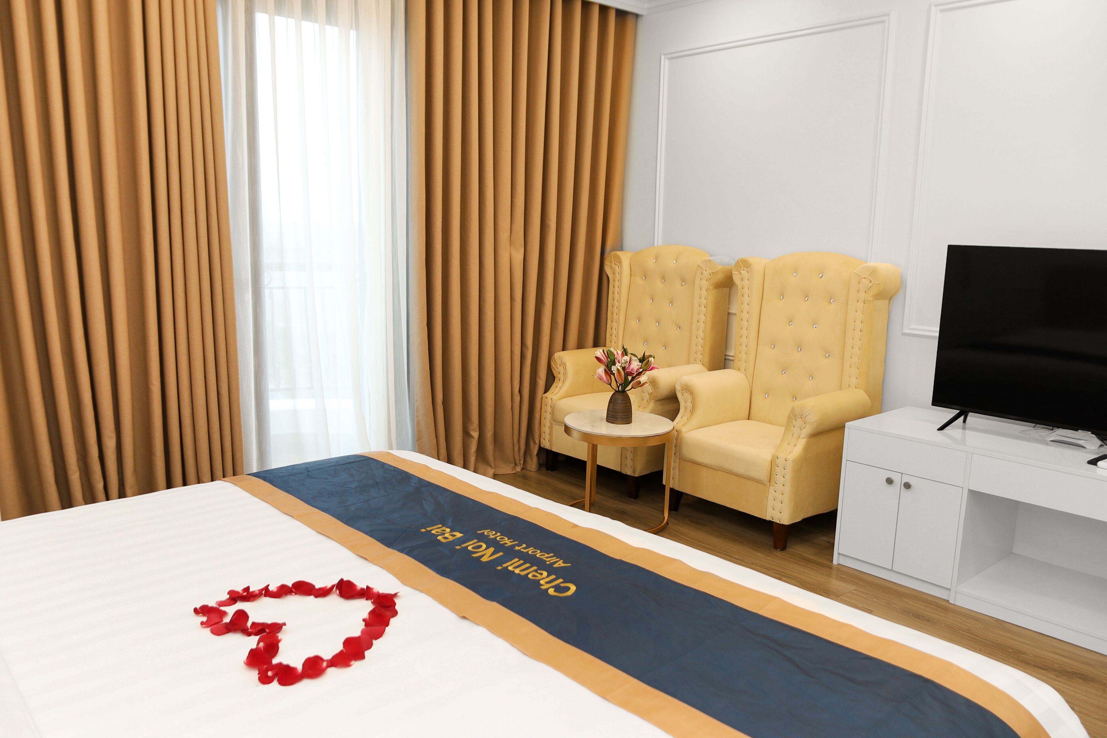 Habitación de hotel con cama, televisor, dos sillones, una mesita con un florero y cortinas.