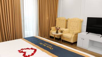 Habitación de hotel con cama, televisor, dos sillones, una mesita con un florero y cortinas.