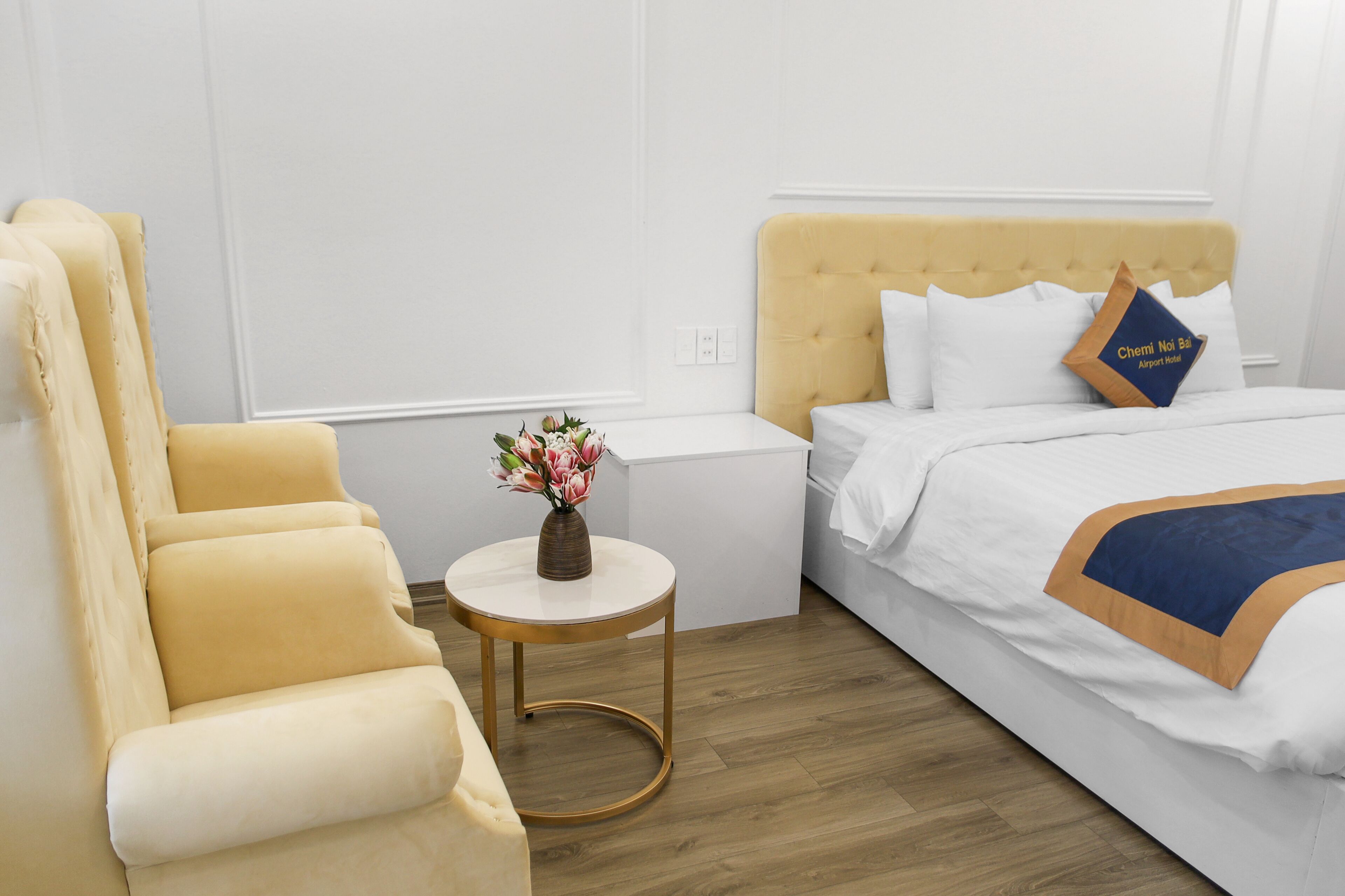 A hotel room with a yellow armchair, a small round table with a vase of flowers, and a bed with white bedding and a blue pillow.