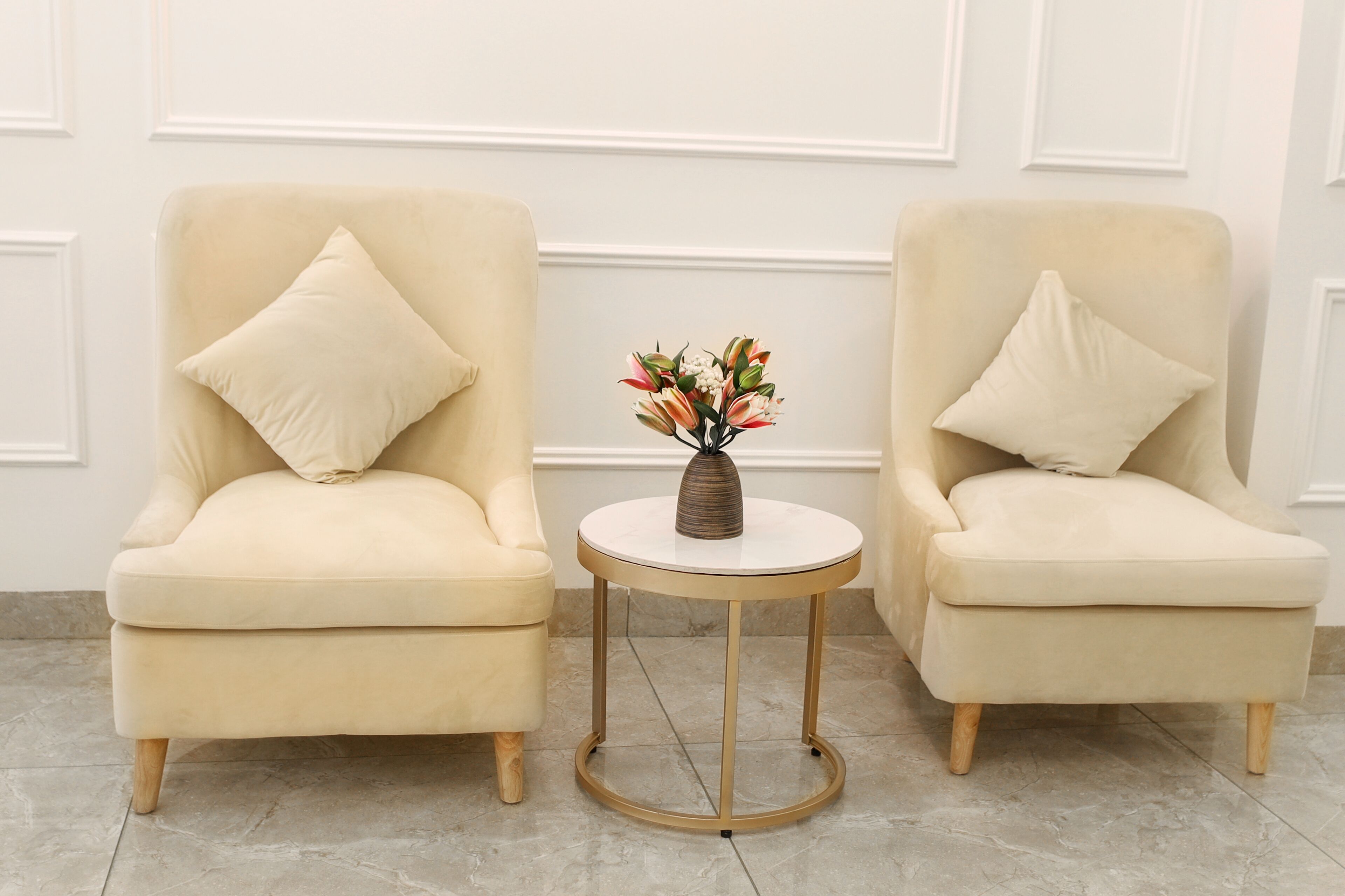 Two cream-colored armchairs with wooden legs and matching cushions, positioned on a tiled floor with a small round table between them.