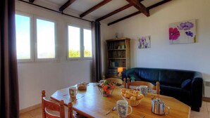 Un comedor con una mesa de madera, sillas y una estantería. Hay ventanas a la izquierda, un sofá a la derecha y un cuadro en la pared.