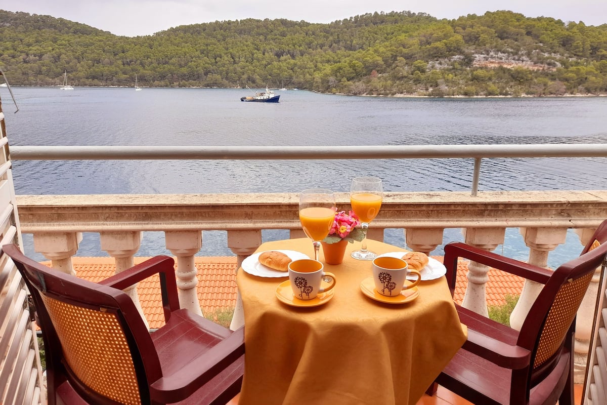 A table with a yellow tablecloth, two chairs, and a view of a lake with boats and a forested hill in the background.