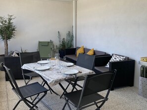 Un coin repas avec une table dressée pour quatre personnes, entourée de chaises noires, d’un canapé gris avec des coussins jaunes et d’une plante en pot.