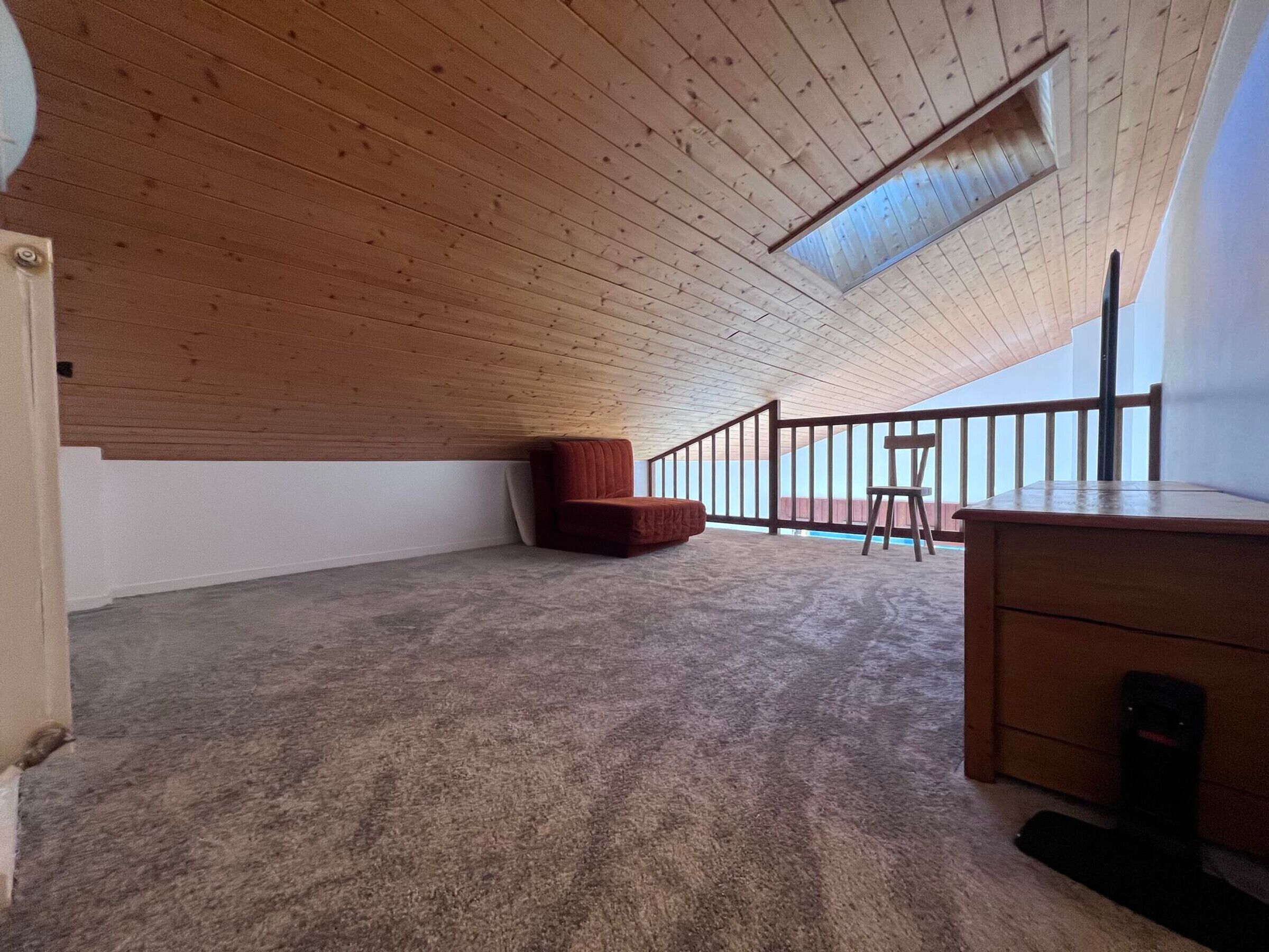 Um quarto de sótão espaçoso com teto inclinado, uma escrivaninha de madeira, uma cadeira vermelha e uma pequena janela.