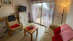 A living room with a sofa, a chair, a small table, a TV on a cabinet, and a sliding glass door with curtains.
