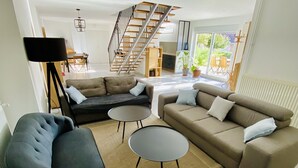 A modern living room with a sectional sofa, two armchairs, a coffee table, and a floor lamp. A staircase leads to an upper level. - Maison Bourgogne 400m2, 2 Piscines Intérieure/extérieure,spa, Commerces à 50m (Gueugnon)