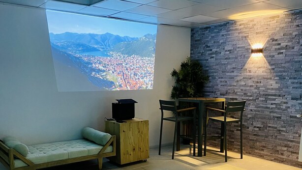 A modern indoor pool area with a projection screen displaying a cityscape, a wooden bench, a table with chairs, and a brick wall with a wall-mounted light fixture. - Maison Bourgogne 400m2, 2 Piscines Intérieure/extérieure,spa, Commerces à 50m (Gueugnon)