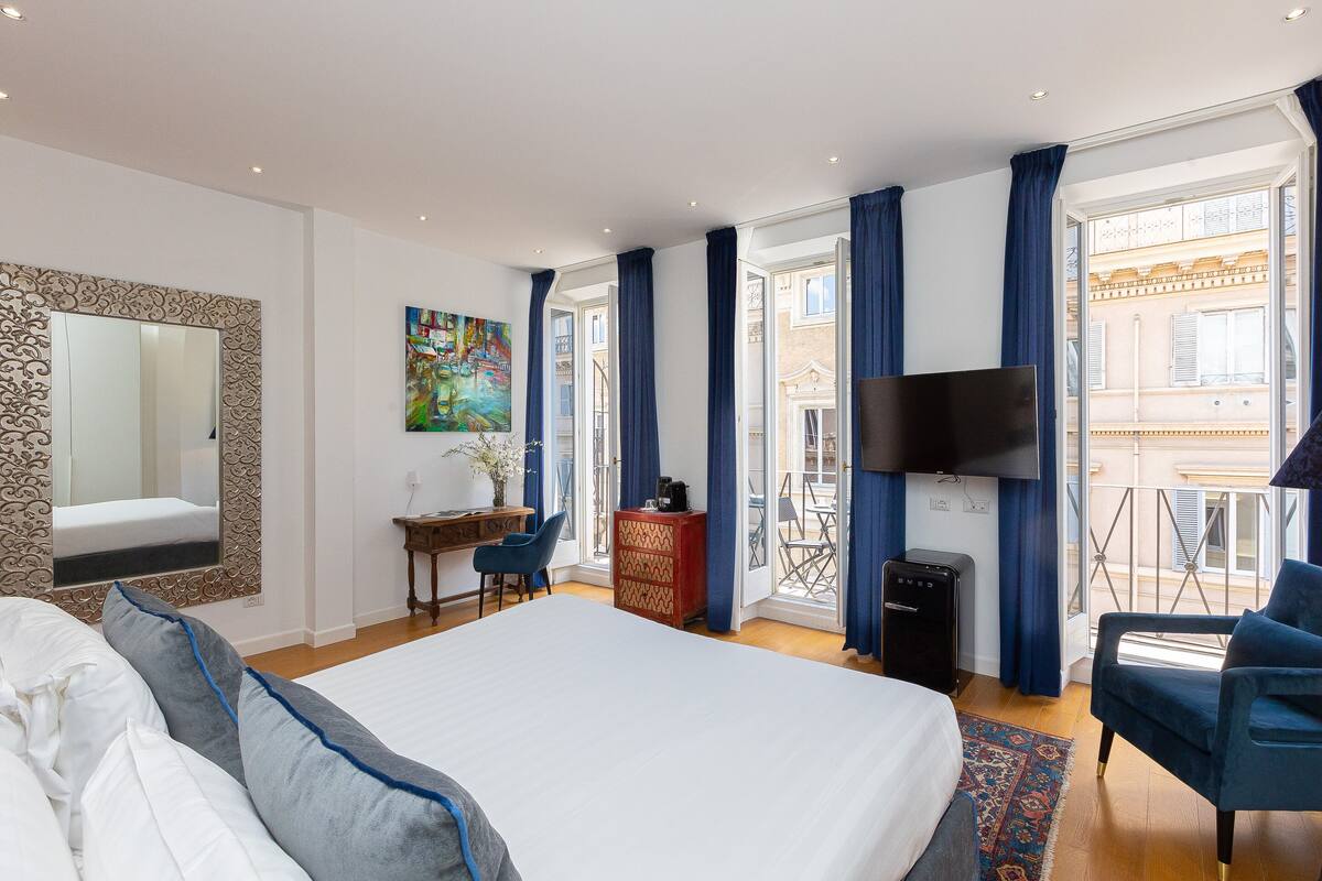 A hotel room with a large bed, a desk with a chair, a TV, and a balcony with a view of the city.