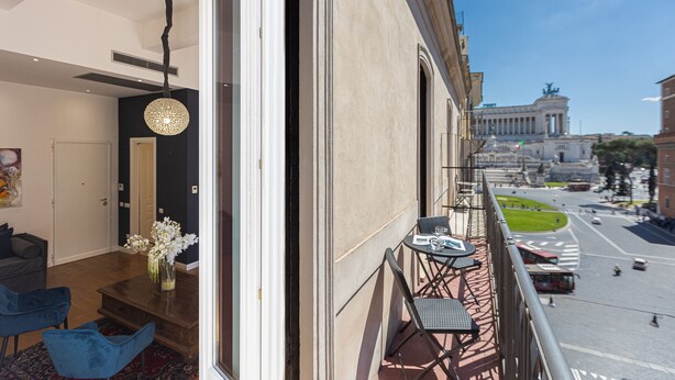 A balcony with a view of a city street, a dining table with chairs, and a vase with flowers. - SUITE DELLA PIGNA (Rome)