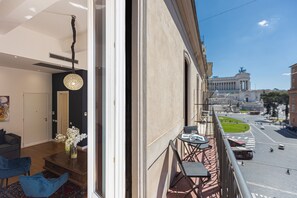 A balcony with a view of a city street, a dining table with chairs, and a vase with flowers. - SUITE DELLA PIGNA (Rome)