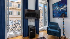 A living room with a TV, a blue armchair, a small black heater, and a balcony with outdoor furniture. - SUITE DELLA PIGNA (Rome)