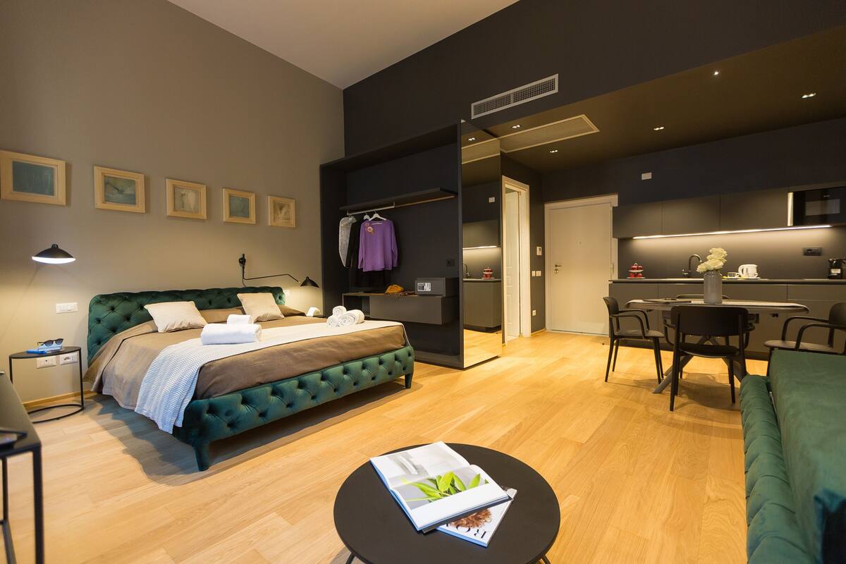 A modern bedroom with a green upholstered bed, a small bedside table, and a nightstand. There is a dining area with a table and chairs, and a kitchen area with a microwave and a sink.