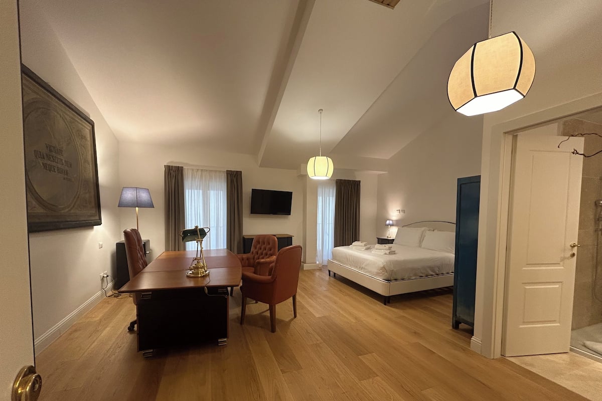 A modern hotel room with a wooden floor, a dining table with chairs, a bed, a TV, and a large window with curtains.