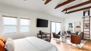 Ein geräumiges Schlafzimmer mit einem großen Bett, einem Schreibtisch, einem Stuhl und einem Sofa. Ein Fernseher ist an der Wand angebracht und es gibt ein Fenster mit Jalousien.