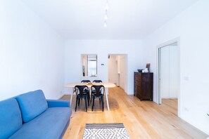 A modern living room with a blue sofa, a dining table with chairs, a dark wooden cabinet, and a patterned rug.