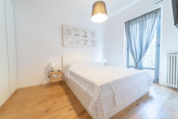 A bedroom with a bed, a bedside table with a lamp, a window with curtains, and a ceiling light fixture.