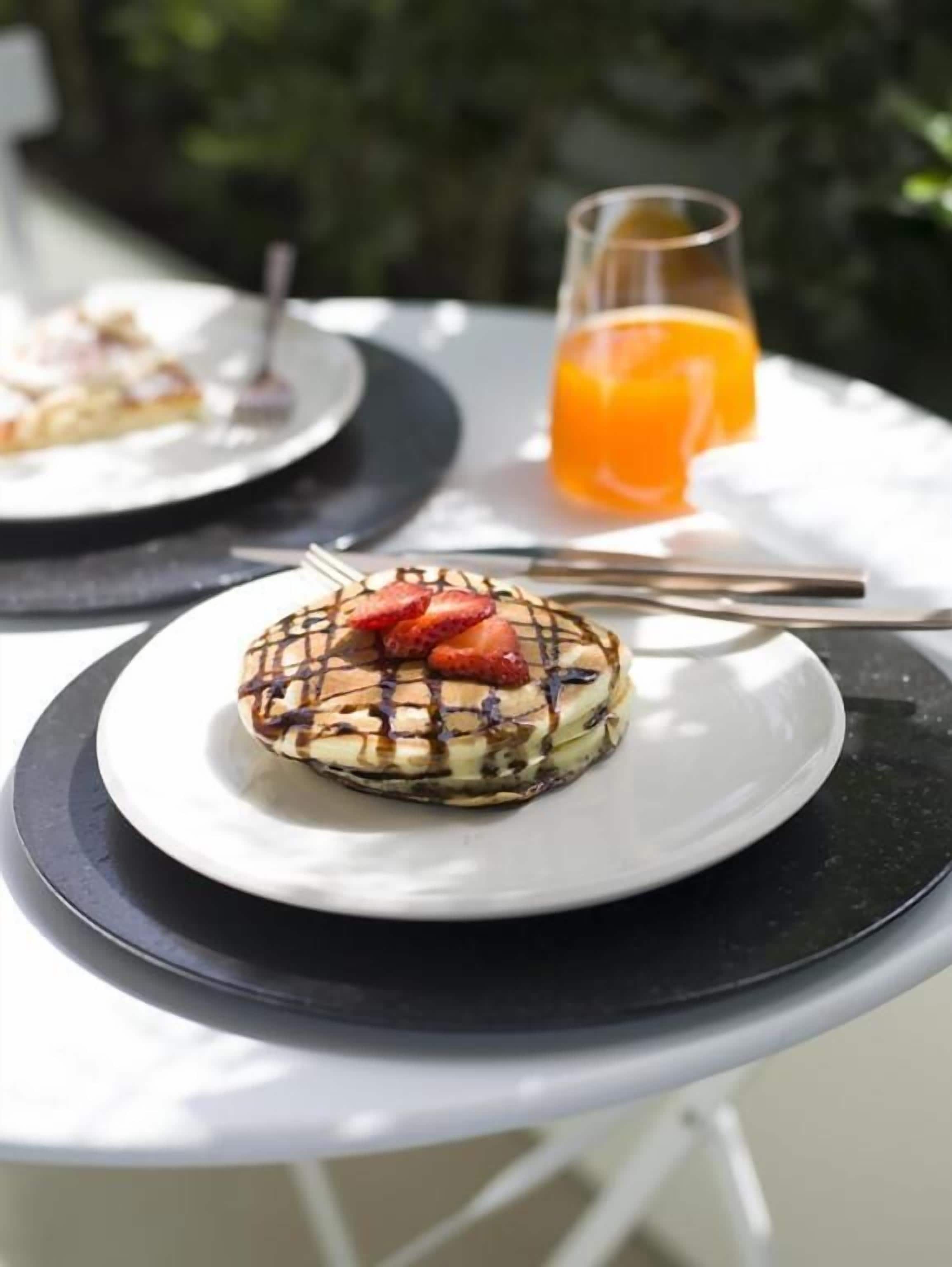 Un dessert servi dans une assiette blanche, nappé de chocolat et agrémenté de fraises, un verre de jus d’orange et une fourchette posée sur la table.