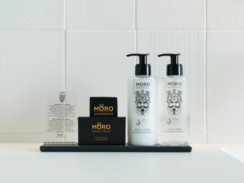 En hylla i badrummet med en svart låda märkt "MORO ACCESSORIES", två flaskor "MORO BODY LOTION" och en svart "MORO Cuffee Cap".