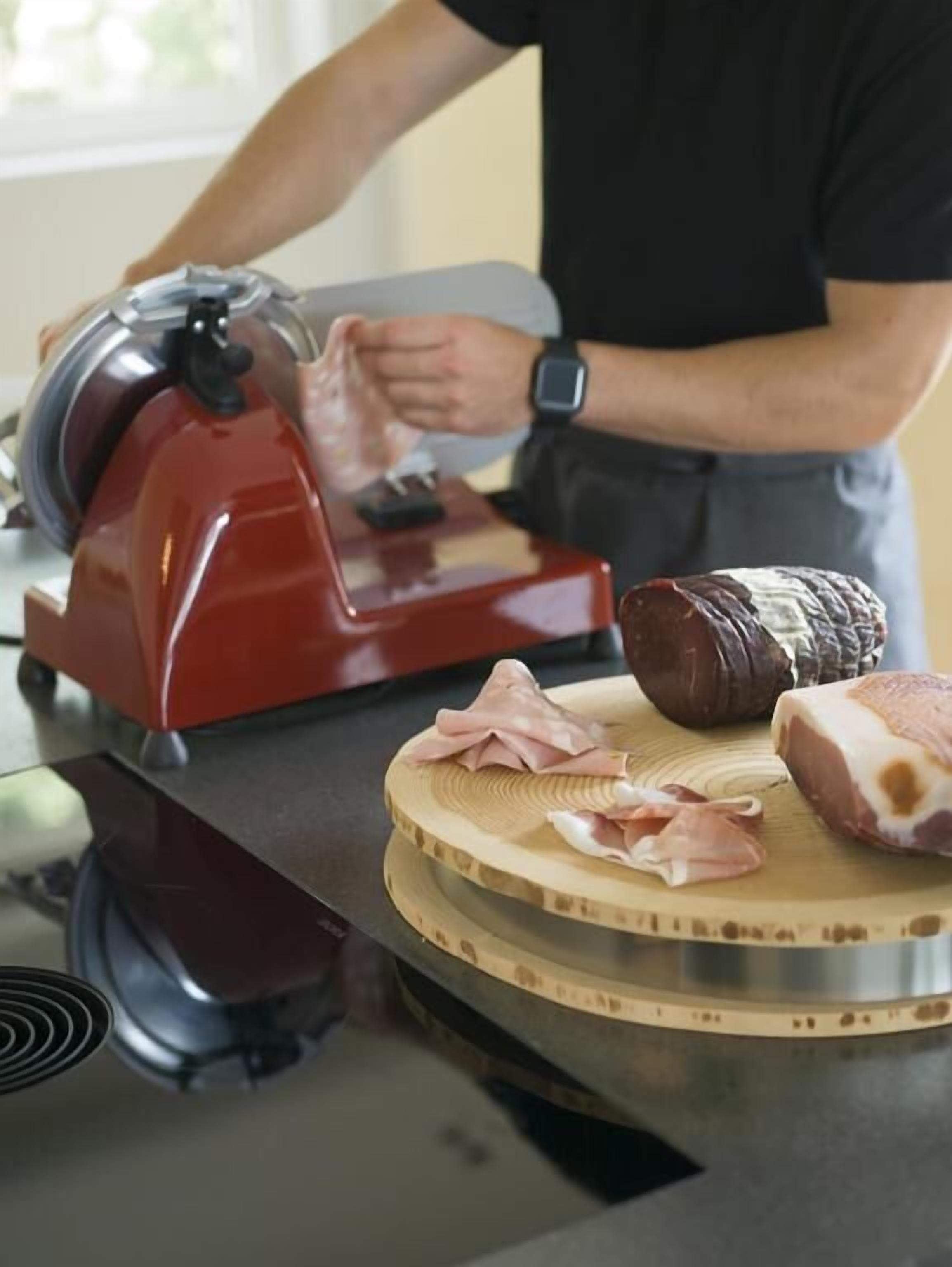 Une personne utilise une trancheuse à viande sur une planche à découper, à côté de morceaux de viande crue et d’une tranche de viande déjà coupée.