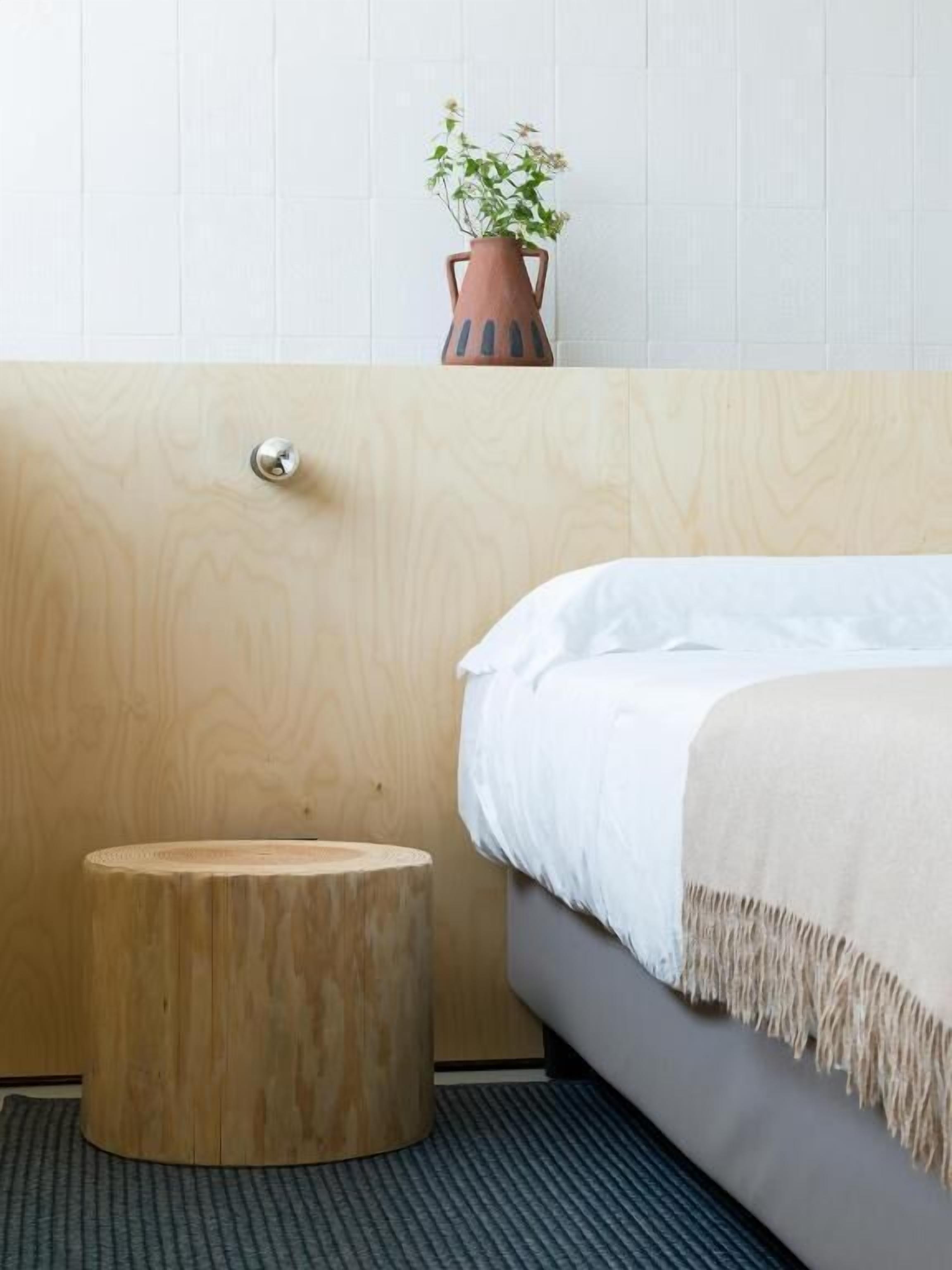 A modern bedroom with a wooden headboard, a round wooden stool, a bed with white bedding, and a potted plant on the headboard.