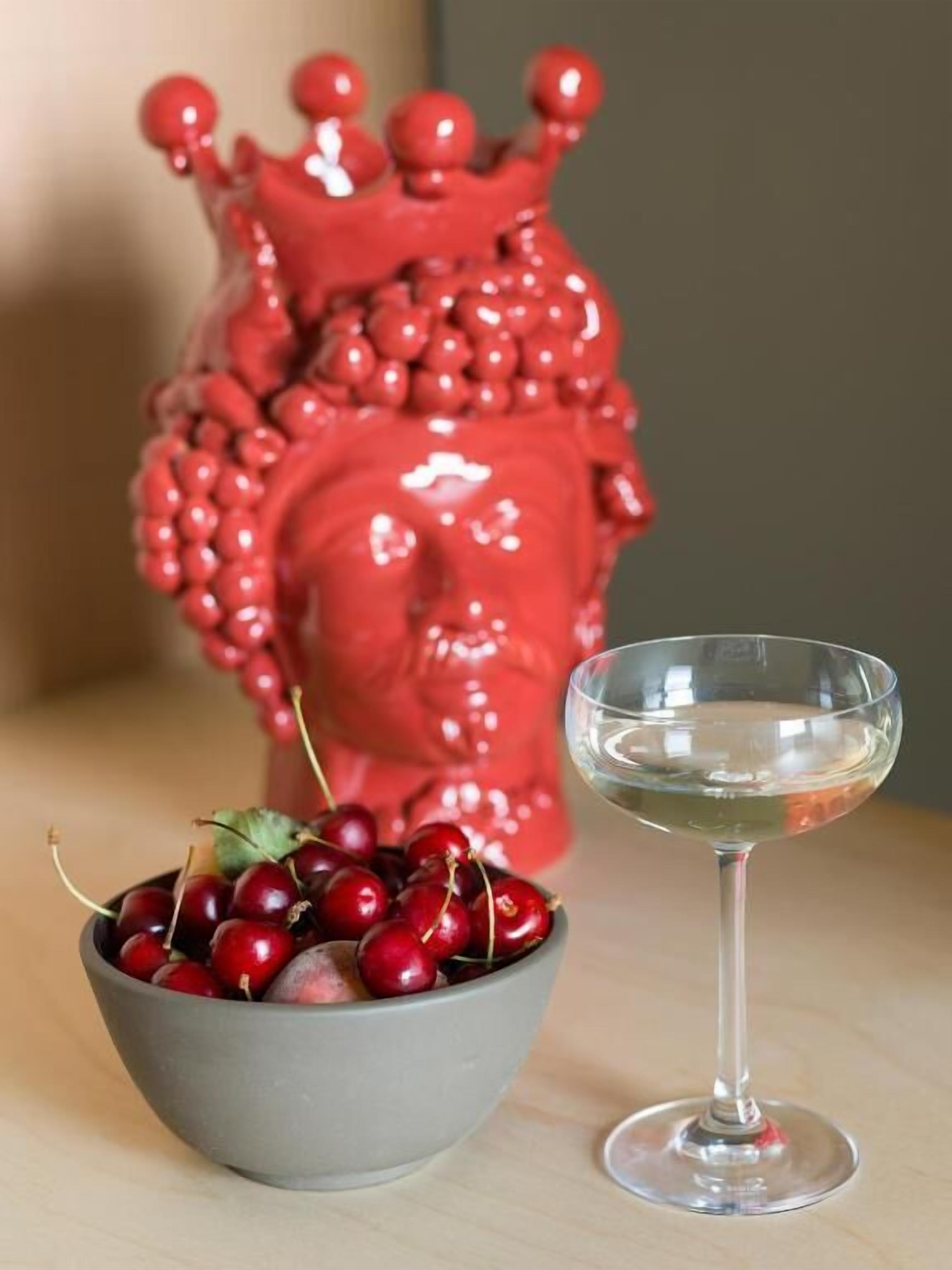 Una ciotola di ciliegie e un bicchiere di vino bianco su un tavolo, con un oggetto a forma di cuore rosso sullo sfondo.