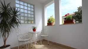 A room with a large potted plant, two white chairs, a small table, and a window with a view of a building and plants outside.