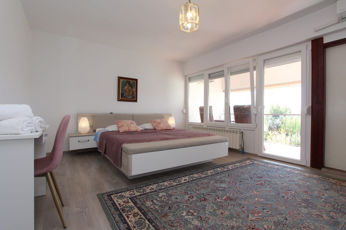 A bedroom with a bed, bedside tables, a chair, a large window, and a view of the outdoors.