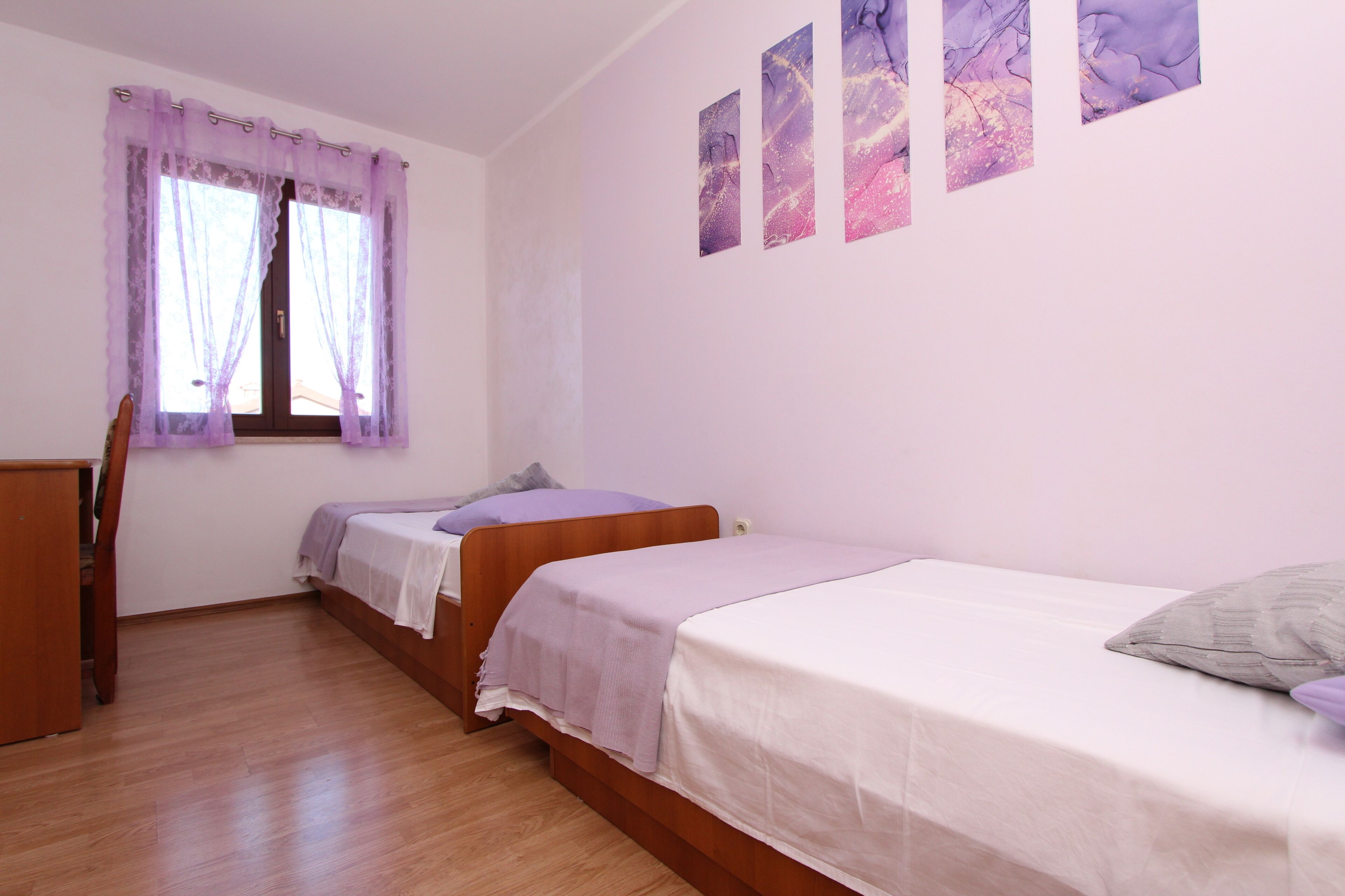 Ein Schlafzimmer mit zwei Betten, einem Holztisch, einem Fenster mit violetten Vorhängen und Wandbildern.