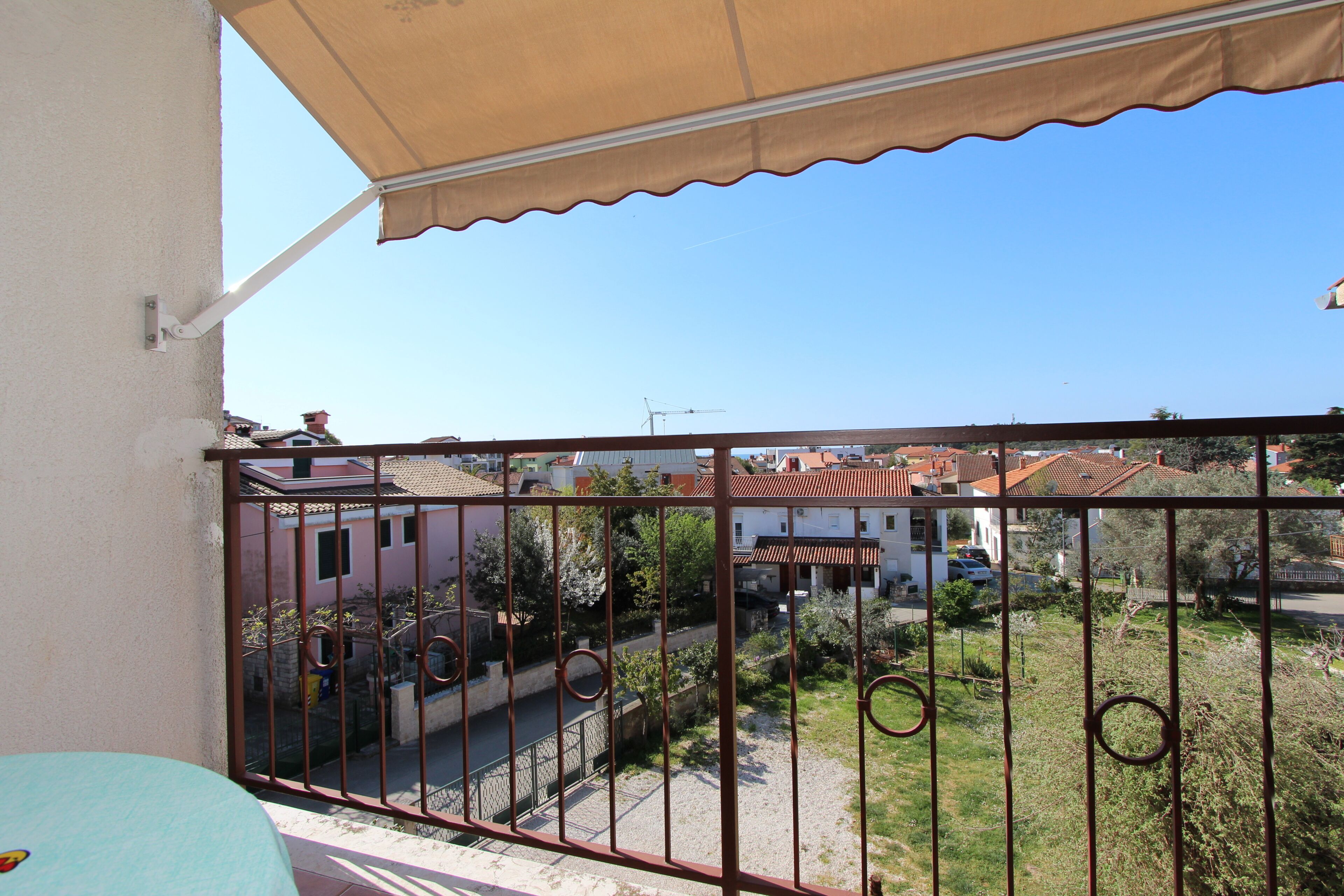 A balcony with a metal railing, a beige awning, and a view of a residential area with houses and trees.