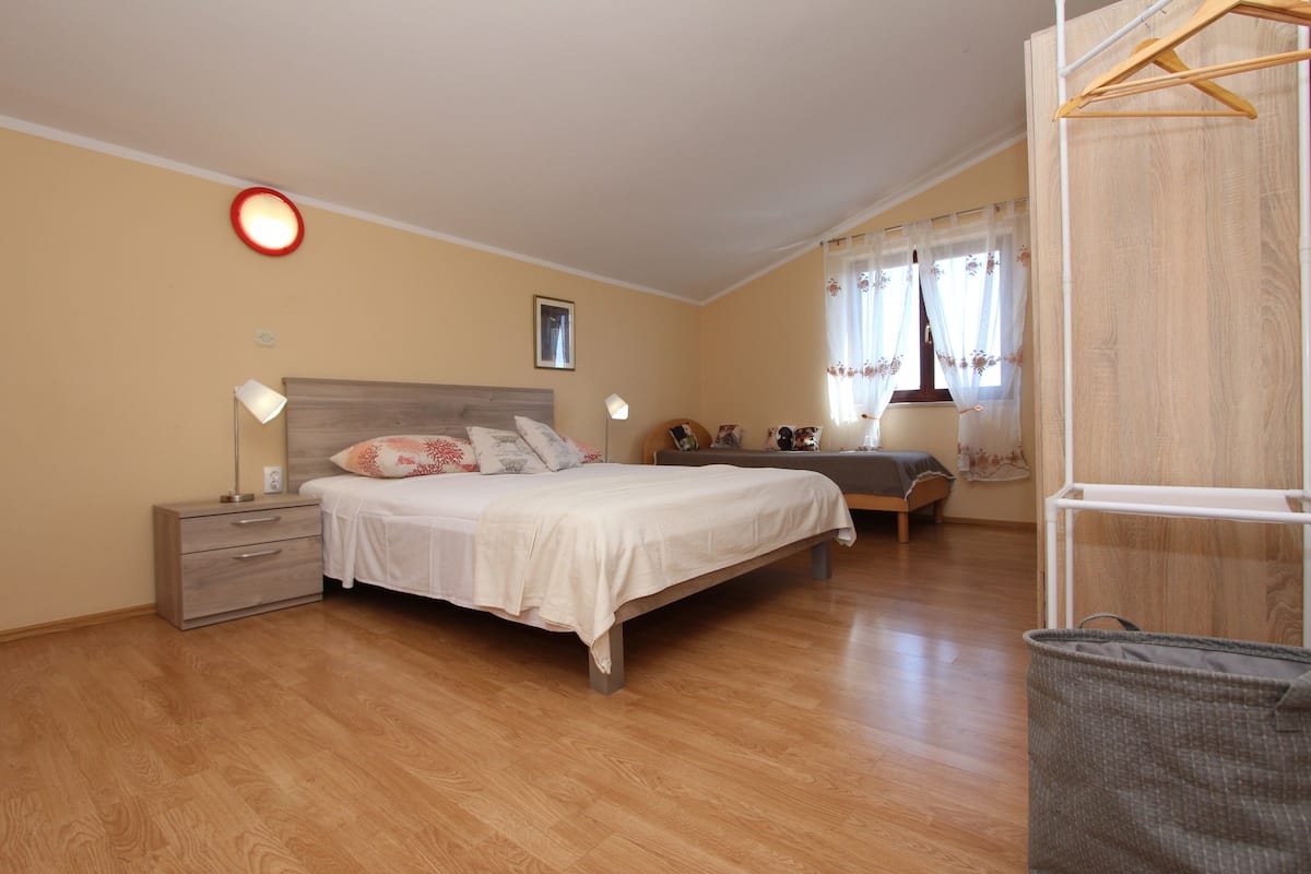 A bedroom with a bed, bedside tables, a lamp, a chair, a window with curtains, and a laundry basket.