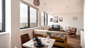 Ein moderner Essbereich mit einem Tisch für zwei Personen, einer Couch mit Decke und Blick auf die Stadt durch große Fenster.