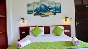 Une chambre d’hôtel avec un lit, une couvre-lit verte, du linge de lit blanc, un tableau représentant des vagues accroché au mur et deux lampes fixées au mur.