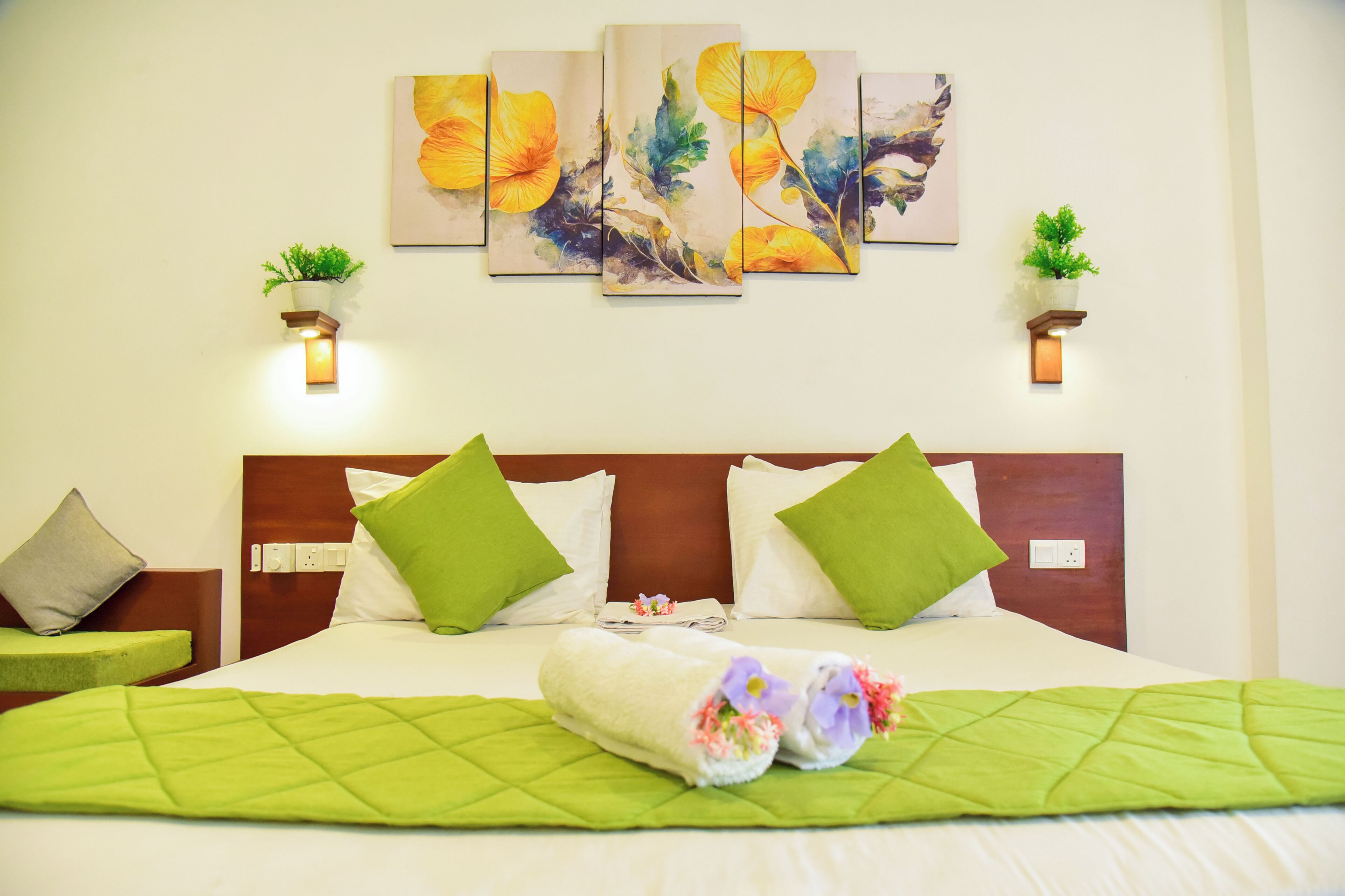 A hotel room with a bed, green pillows, a green quilt, artwork on the wall, and wall-mounted plants.